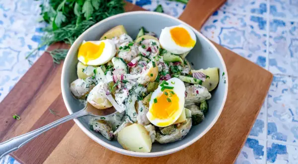 Schüssel mit cremigem Kartoffelsalat mit Gurken, Zwiebeln und gekochten Eiern, garniert mit frischen Kräutern.
