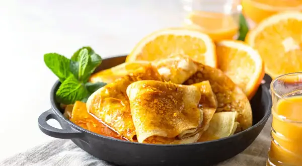 Złociste crepes suzette w sosie pomarańczowym, udekorowane miętą i plasterkami pomarańczy, obok szklanki soku.