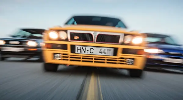Żółta Lancia Delta Integrale w centrum, ścigana przez niebieski i czarny samochód.