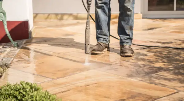 Un homme nettoie sa terrasse carrelage avec un nettoyeur haute pression. L'eau éclabousse les dalles.