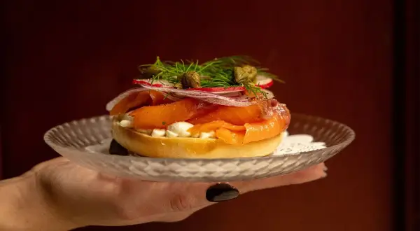 En hand håller upp en bagel med rökt lax, färskost, rödlök, rädisor och kapris. Perfekt för en lyxig bank hotel middag.