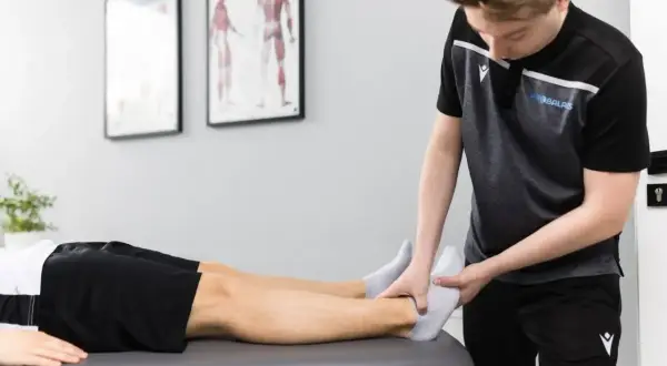 Fizjoterapeuta w sporcie delikatnie bada stopę zawodnika leżącego na kozetce.