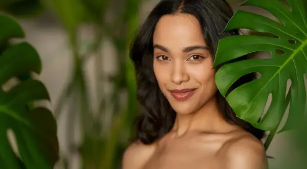 Mujer con maquillaje efecto bronceado, sonriendo entre hojas verdes.