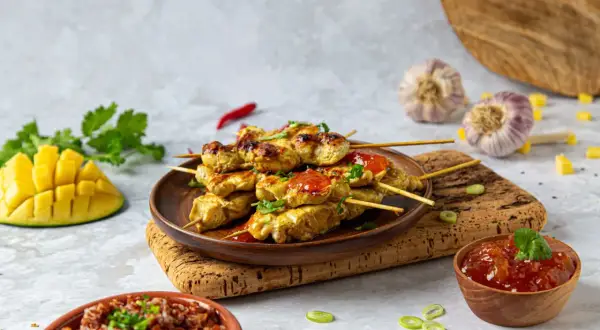 Aksamitne satay z kurczaka na patyczkach, polane sosem, obok ryż, mango i czosnek.