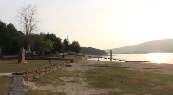 Un tranquilo atardecer en la playa fluvial o corgo, con un muelle y vegetación verde.