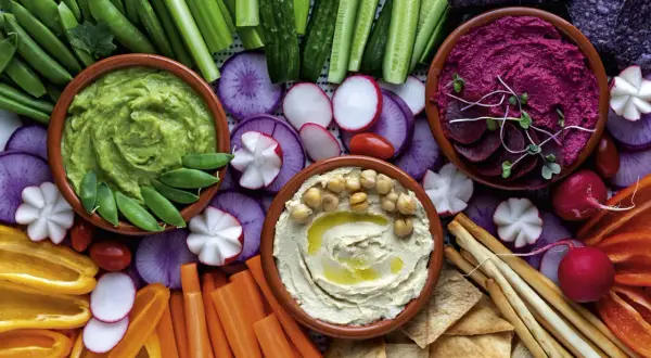 Bunte Auswahl an Dip-Rezepten: cremiger Hummus, grüner Avocado-Dip und Rote-Bete-Dip mit frischem Gemüse und Brot.