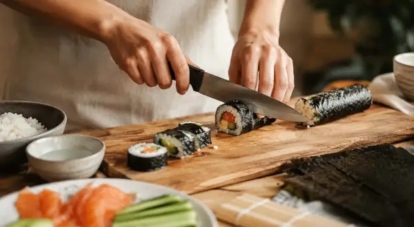 Ręce kroją rolkę sushi na desce. Widać składniki: ryż, łososia, ogórka. Pyszne domowe sushi w przygotowaniu.