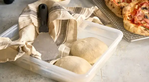 Pizzadeg redo att baka ut. En degskrapa och en kökshandduk ligger bredvid en plastlåda med två degbollar.
