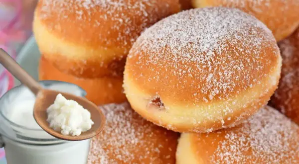 Puszyste pączki z lukrem, posypane cukrem pudrem, obok słoiczka z twarożkiem i drewnianej łyżeczki.