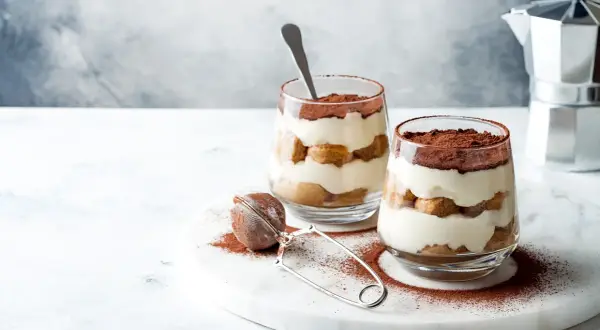 Dwa pucharki z tiramisu bez jajek, przełożone biszkoptami i kremem, posypane kakao. Obok sitko do kakao i kawiarka.