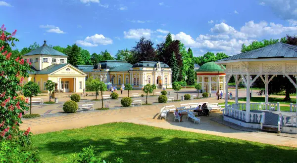 Ein sonniger Tag in einer **Therme Tschechien**. Gelbe Kurgebäude mit grünen Dächern, ein weißer Pavillon und gepflegte Gärten laden zum Entspannen ein.