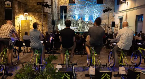 Spettatori pedalano su cyclette per alimentare un'esibizione di teatro a pedali in una piazza illuminata.