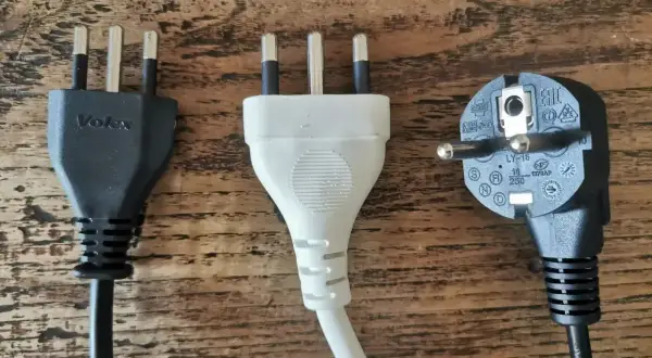 Trzy wtyczki elektryczne, w tym dwie z trzema bolcami, typowe dla gniazdek we Włoszech, leżą na drewnianym blacie.