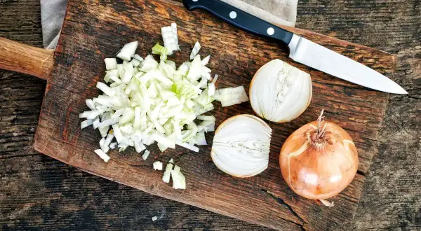 Cipolla cruda digeribile, tagliata finemente su un tagliere di legno, accanto a una cipolla intera e a metà. Un coltello è pronto all'uso.