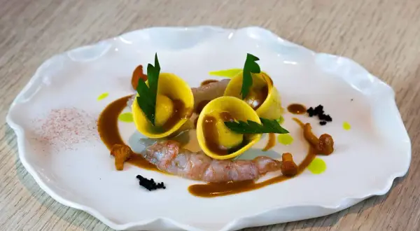 Trois raviolis italiens dorés, garniture de crevettes, champignons et sauce brune sur une assiette blanche ondulée.