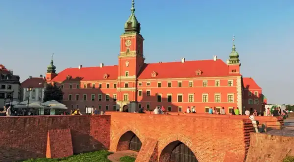 Zamek Królewski w Warszawie, z czerwoną fasadą i zieloną wieżą, widziany zza ceglanego mostu.