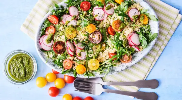 En stor bulgursallad med körsbärstomater, rädisor och ruccola, toppad med en grön dressing. Serveras med en skål pesto och extra tomater.