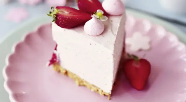 Ein Stück Erdbeer Cheesecake mit frischen Erdbeeren und Baiser-Tupfern auf einem rosa Teller.