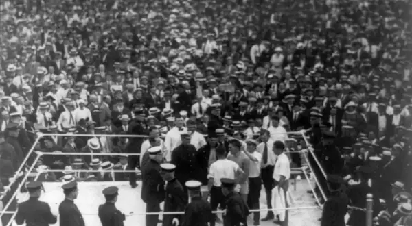 Boxing ring with fighters and officials, surrounded by a massive, cheering crowd in vintage attire.