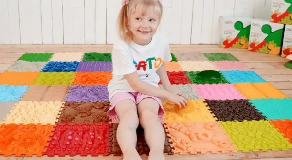 Uśmiechnięta dziewczynka bawi się na kolorowej macie sensorycznej dla niemowląt, odkrywając różne faktury i kształty.