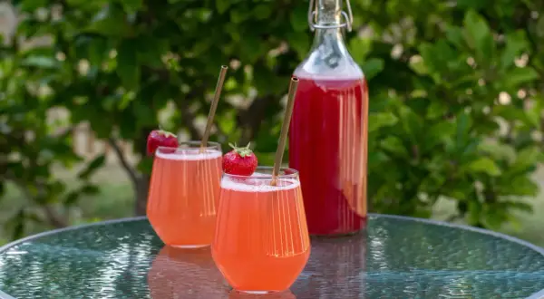 Två glas med rosa rabarbersaft, garnerade med jordgubbar och sugrör, står på ett glasbord. En flaska med saft i bakgrunden. Perfekt för ett rabarbersaft recept.