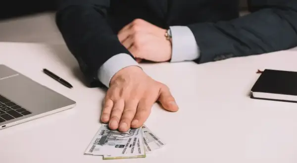 Dłoń mężczyzny w garniturze podaje banknoty rubli na białym biurku obok laptopa.