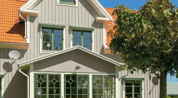 Vitt hus med gråa knutar och orange tak. Gröna fönster och en stor lönn skänker skugga.