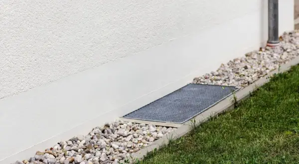 A gravel bed and grate next to a white wall, showing how to level ground for drainage.