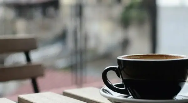 En svart kopp med lungo kaffe står på ett träbord. I bakgrunden syns en bänk och en suddig stadsmiljö.