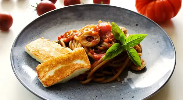 Makaron z halloumi na talerzu, z sosem pomidorowym, oliwkami i bazylią. Obok leżą świeże pomidory.