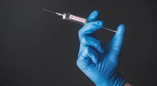 Ręka w niebieskiej rękawiczce trzyma strzykawkę z napisem VACCINE. To symbol szczepień, także w Tunezji.