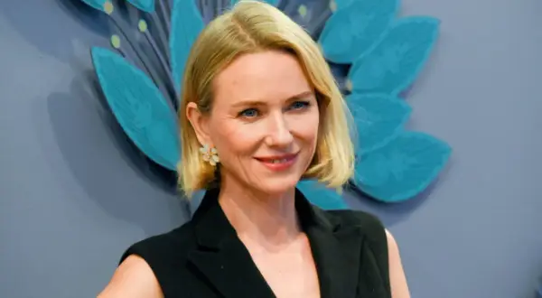 Uśmiechnięta Naomi Watts, znana z wielu filmów, w czarnej stylizacji i z kwiatową broszką.