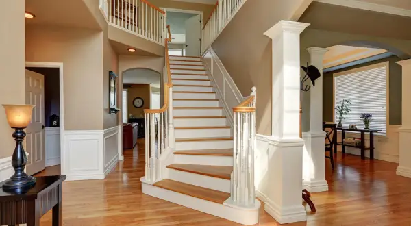 A grand foyer with a sweeping staircase featuring white painted risers and wooden treads. The staircase leads to an upper landing, with a view into a living area.
