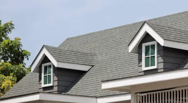 A close-up of a house roof showcasing different types of shingles in a grey hue, with dormer windows peeking out.