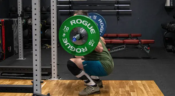 Mężczyzna wykonuje głęboki **squat** z ciężarem na plecach w siłowni. Zielone talerze Rogue ważą 10 kg każdy.