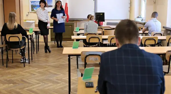 Uczniowie siedzą w ławkach, czekając na egzamin. Nauczycielki stoją z przodu, trzymając dokumenty. Ile trzeba mieć procent żeby zdać egzamin zawodowy?