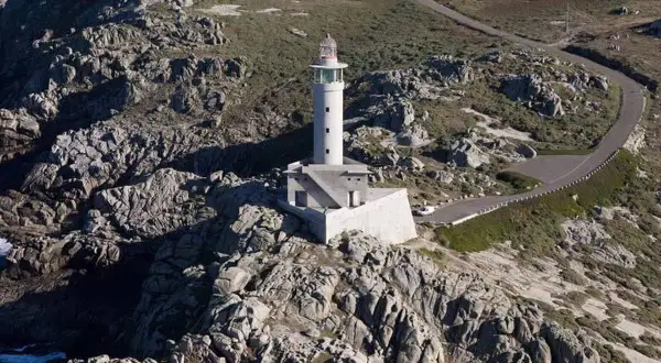 Faro blanco en una punta rocosa, con una carretera serpenteante que sube hacia él.