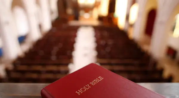 Czerwona Biblia leży na pierwszym planie, w tle widać rzędy pustych ławek w kościele. Szukasz odpowiedzi, jak przejść na protestantyzm?