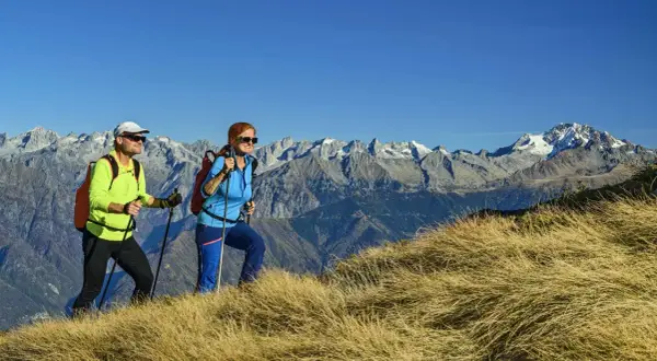 Zwei Wanderer genießen die Aussicht auf die Alpen. Sie gehen auf einem grasbewachsenen Hang, Stöcke in den Händen, mit schneebedeckten Gipfeln im Hintergrund.