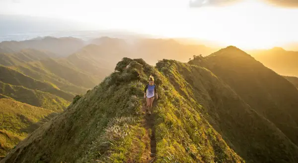 Une femme court sur un sentier de montagne verdoyant au coucher du soleil, profitant d'un parcours spectaculaire.