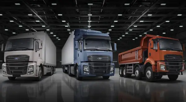 Trois camions Ford F-Line : un blanc avec remorque, un bleu avec remorque, et un orange benne basculante, alignés dans un entrepôt.