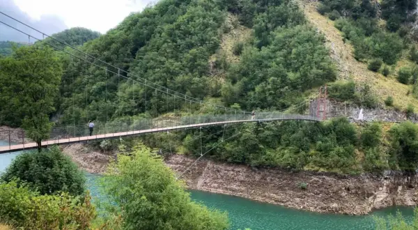 Un ponte sospeso attraversa un lago turchese, circondato da montagne boscose. Due persone passeggiano sul ponte, godendosi il panorama dei vagli di sotto.