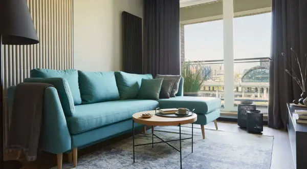 Turkusowa sofa z poduszkami i kocem, niski stolik kawowy z filiżanką kawy. Idealna wysokość ławy do salonu.