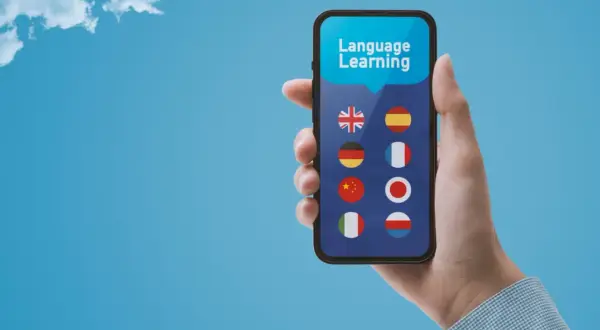 Dłoń trzyma smartfon z aplikacją do nauki języków. Widoczne flagi: Wielkiej Brytanii, Hiszpanii, Niemiec, Francji, Chin, Japonii, Włoch i Polski. Bussu ułatwia podróże.