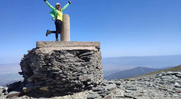 Mujer con el chullo puesto, brazos en alto, celebra en la cima de una montaña rocosa bajo un cielo azul despejado.