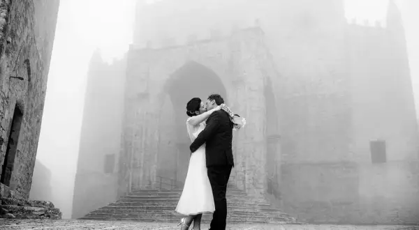 Sposi si baciano davanti a un antico arco in pietra, immersi nella nebbia. Dettagli matrimonio suggestivi.