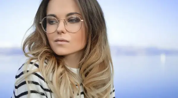 Blondynka w okularach o okrągłych oprawkach, w bluzie w paski, na tle błękitnego nieba i wody.