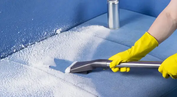 Mani con guanti gialli eseguono il lavaggio divano a secco con schiuma e aspiratore su tessuto blu.