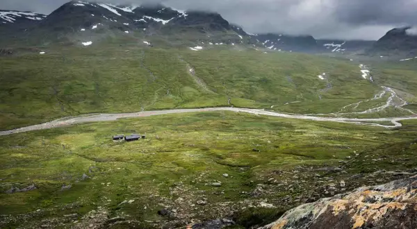 Grönskande fjällandskap med en slingrande älv och små stugor. En del av Kungsleden vid Hemavan, med snö på topparna.