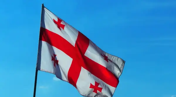 Flaga Gruzji powiewa na tle błękitnego nieba. Biały materiał ozdobiony jest dużym czerwonym krzyżem i czterema mniejszymi krzyżami w rogach.